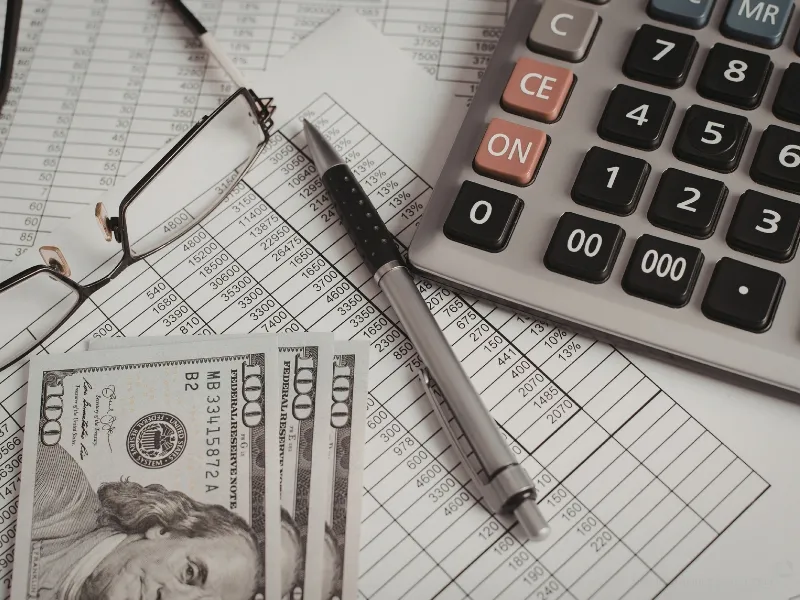 100 dollar bills, glasses, pen, calculator, and a general ledger in accounting