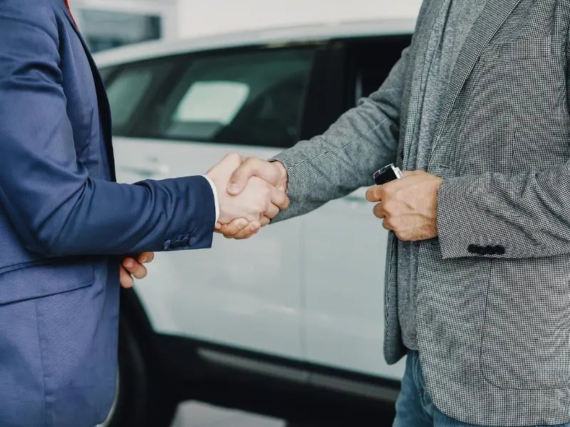 two business men shake hands in the foreground of a car. Image highlights irs business mileage rate