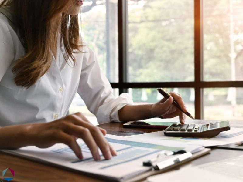 A woman is calculating finances using a calculator and a budget spreadsheet. This featured image is for the article about business debt consolidation.