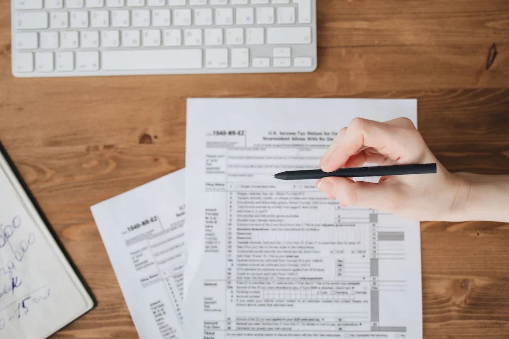 Close-up of a hand holding a pen over US tax forms on a wooden table. Image complements the blog titled Multi State Tax Filing.
